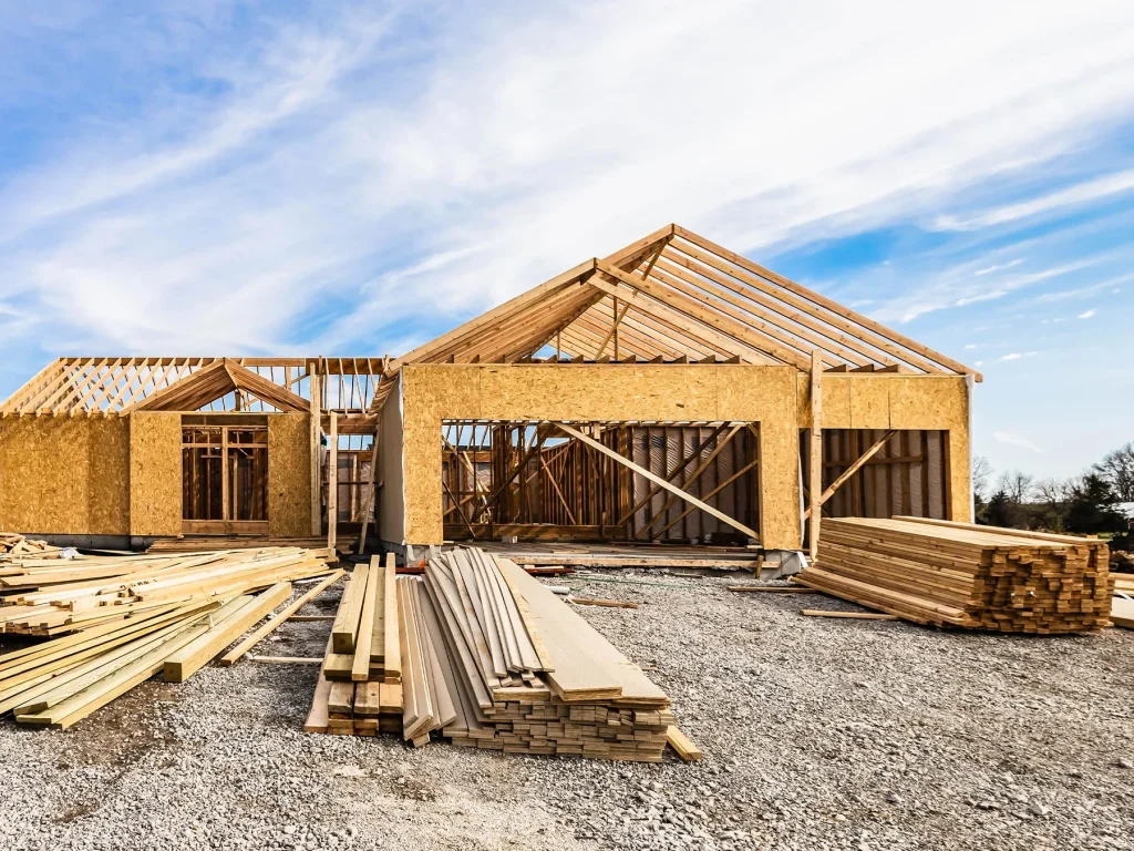 Timber-frame house under construction with roof trusses in place and stacks of timber on a gravel site